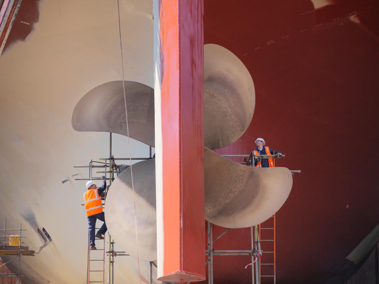 To arbeidere med hjelp og sikkerhetsvest vedlikeholder en stor båtpropell fra stiger