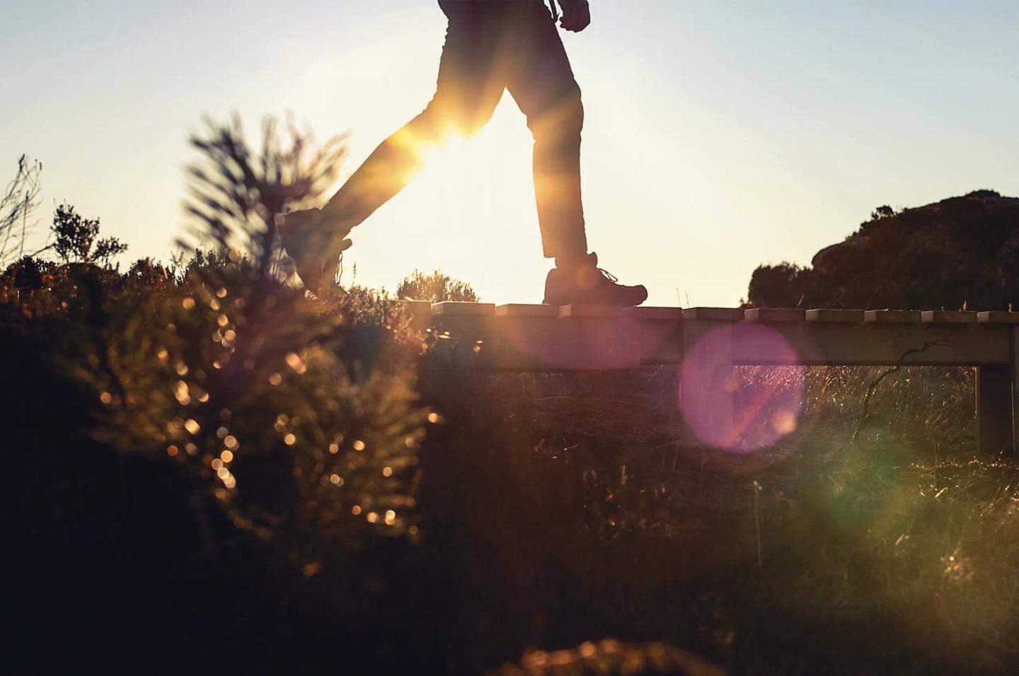 Person i fritidsklær går tur i åpen landskap, og krysser en bekk over en enkel trebro.