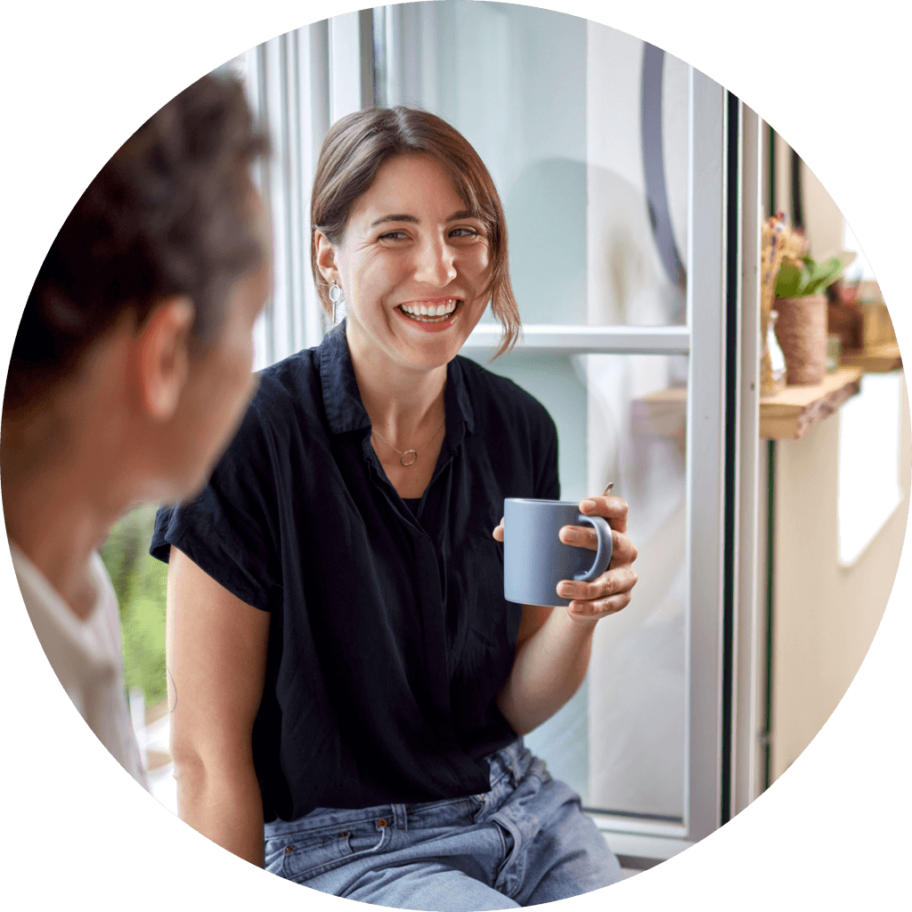Person som sitter i vinduskarm med kaffekopp i hånda og ler til en kollega