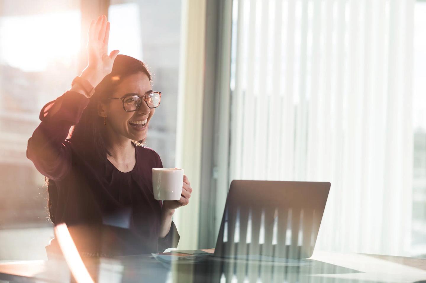Smilende person med på jobb med laptop og kaffekopp løfter hånden for å hilse en kollega velkommen.