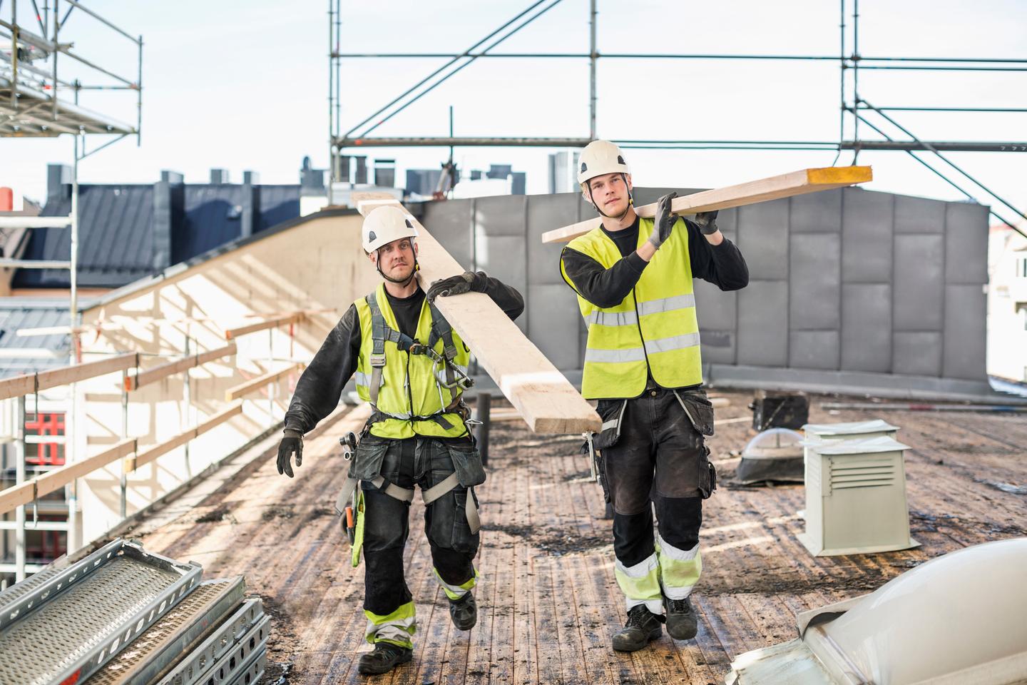 To byggarbeidere på et tak som bærer bjelker iført verneutstyr